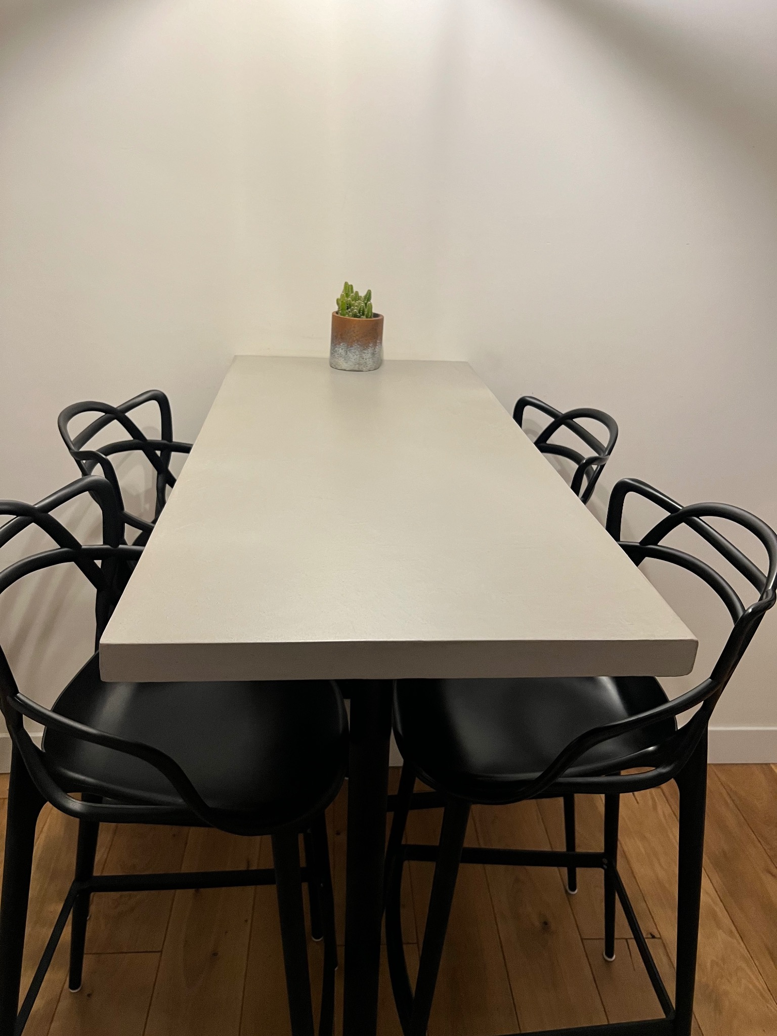Stół z blatem o jednolitej, jasnoszarej powierzchni, otoczony czterema czarnymi krzesłami o nowoczesnym designie, z małą rośliną doniczkową na środku, na drewnianej podłodze i tle jasnej ściany.