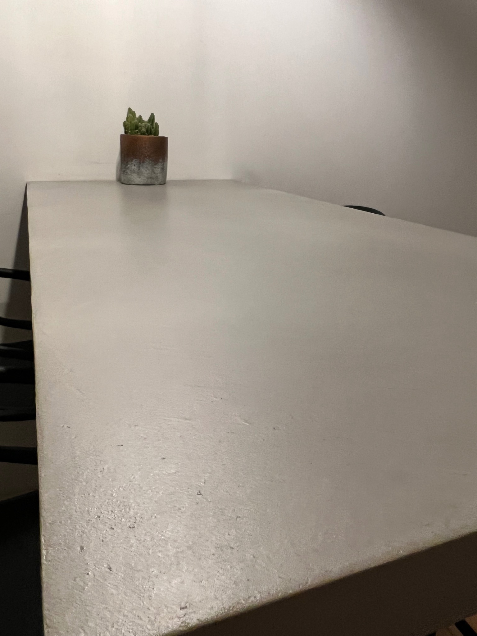 Blat stołu z szarego betonu architektonicznego z delikatną fakturą, na którym stoi mały kaktus w doniczce z efektem ombre, na tle białej ściany.