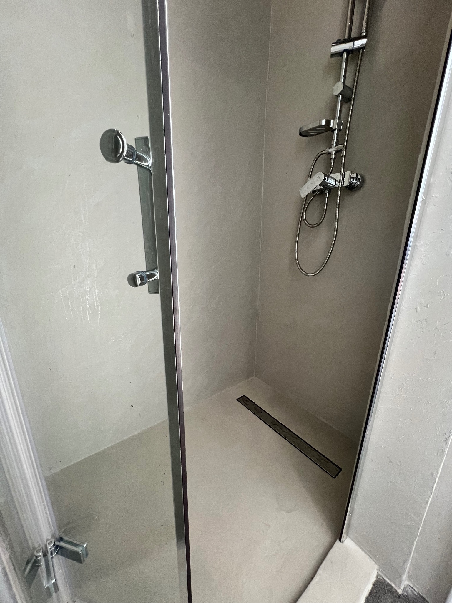 Wnętrze kabiny prysznicowej z odpływem liniowym, ściany wykończone w jasnym odcieniu szarości, bateria prysznicowa z deszczownicą i słuchawką, widoczne detale chromowane.