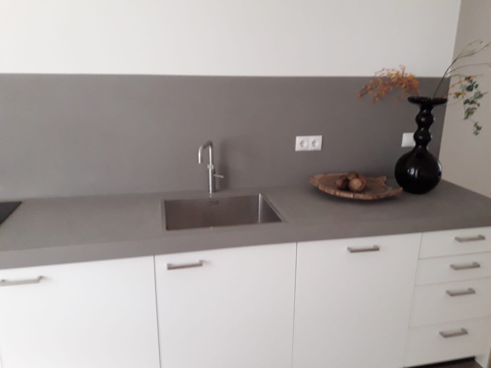Szary blat kuchenny z minimalistycznym zlewem, białe szafki z podłużnymi uchwytami, dekoracyjna czarna waza i drewniana misa z orzechami, szara ściana nad blatem, gniazdka elektryczne.