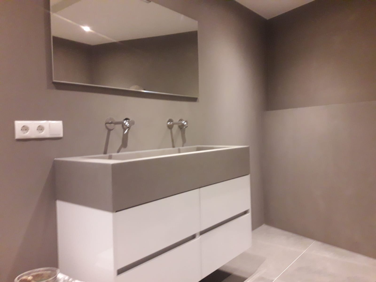 Nowoczesna łazienka z podwójną umywalką z szarą, betonową obudową i białymi szafkami, minimalistyczne baterie, lustro bez ramy, gładkie ściany w odcieniach szarości, widoczne gniazdko elektryczne.