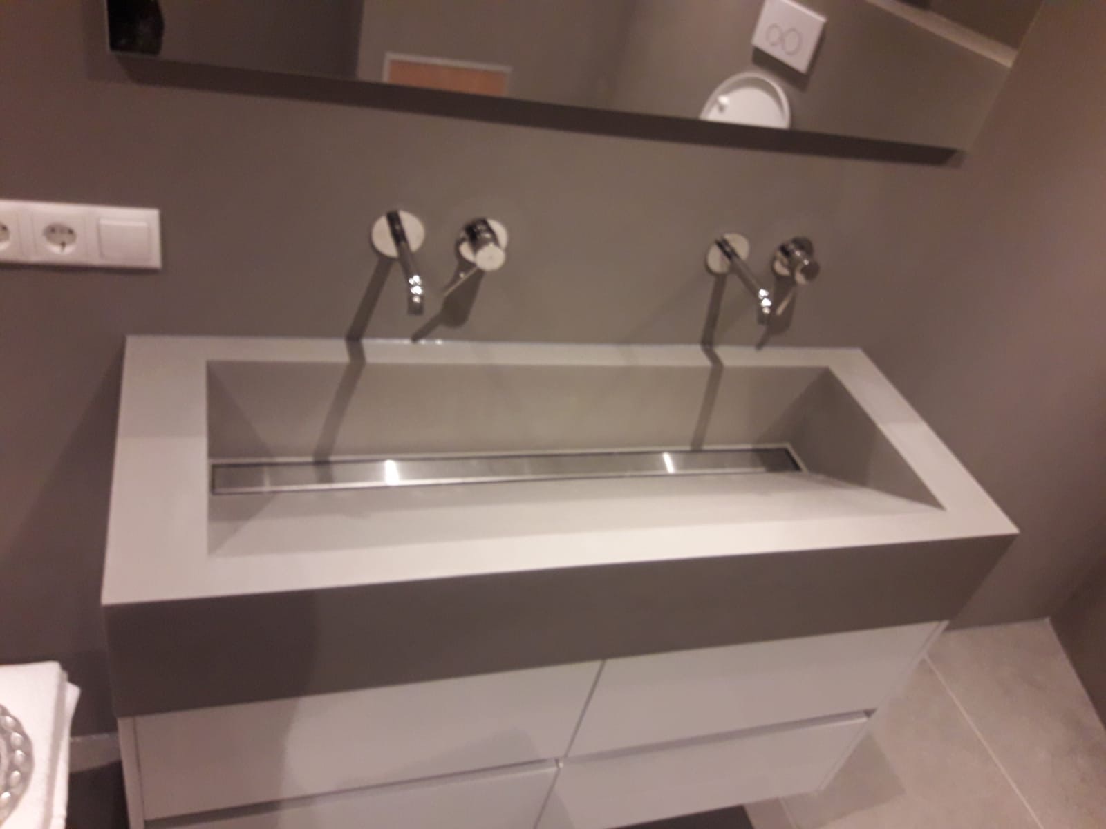 Szara, prostokątna umywalka z dwoma chromowanymi kranami, zamontowana na szafce z szufladami, widoczna w łazience z szarymi ścianami i lustrem.