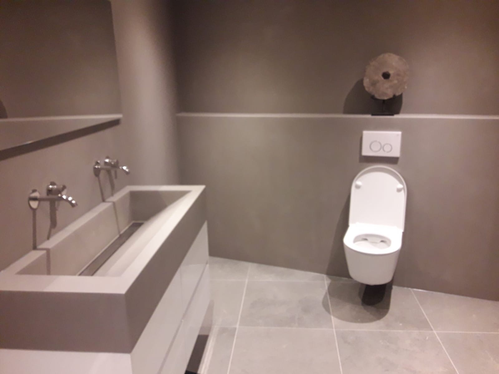 Nowoczesna łazienka w odcieniach szarości z podwójną umywalką o geometrycznych kształtach, chromowanymi bateriami, wiszącą toaletą i dekoracyjnym elementem na ścianie.