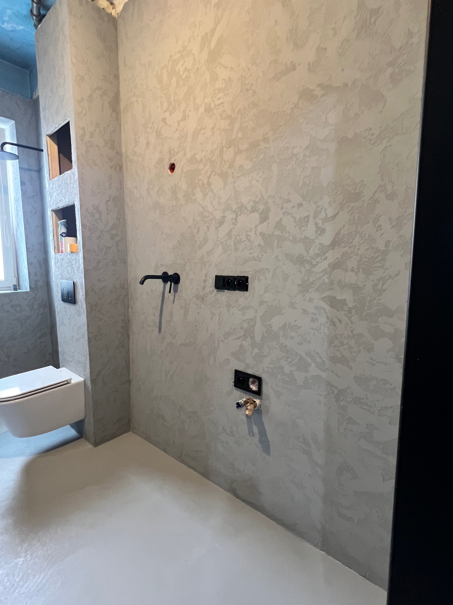 Nowoczesna łazienka z szarą, teksturowaną ścianą, wiszącą toaletą i czarną armaturą, z ukrytymi półkami na kosmetyki i gładką podłogą.