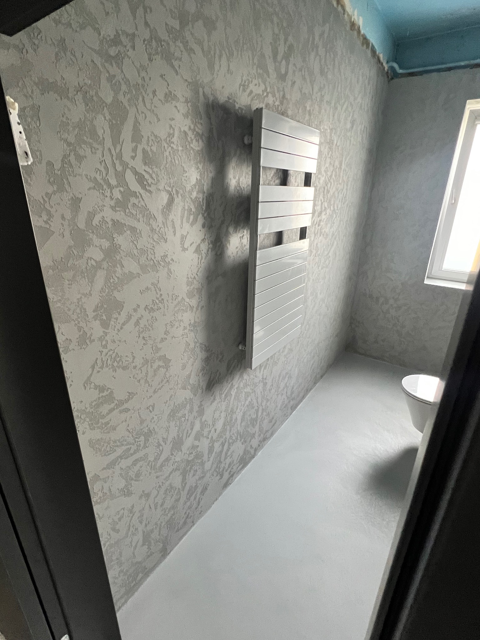 Ściana w łazience z dekoracyjną szarą fakturą, zamontowany biały grzejnik drabinkowy, widoczny fragment toalety i okna, sufit pomalowany na niebiesko.