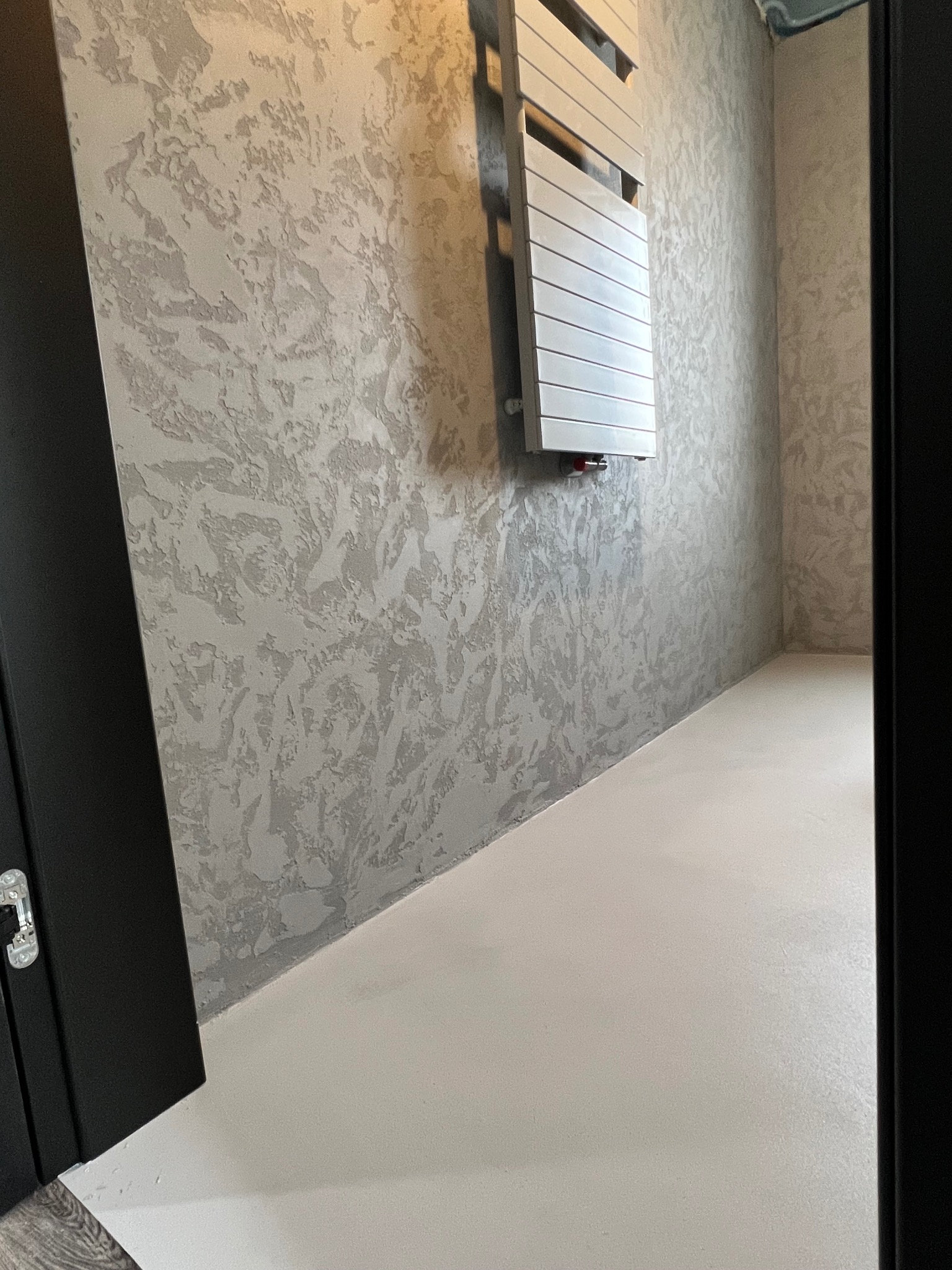 Ściana w łazience z dekoracyjną strukturą w odcieniach szarości, grzejnik drabinkowy na ścianie, biała podłoga, widok zza otwartych czarnych drzwi.