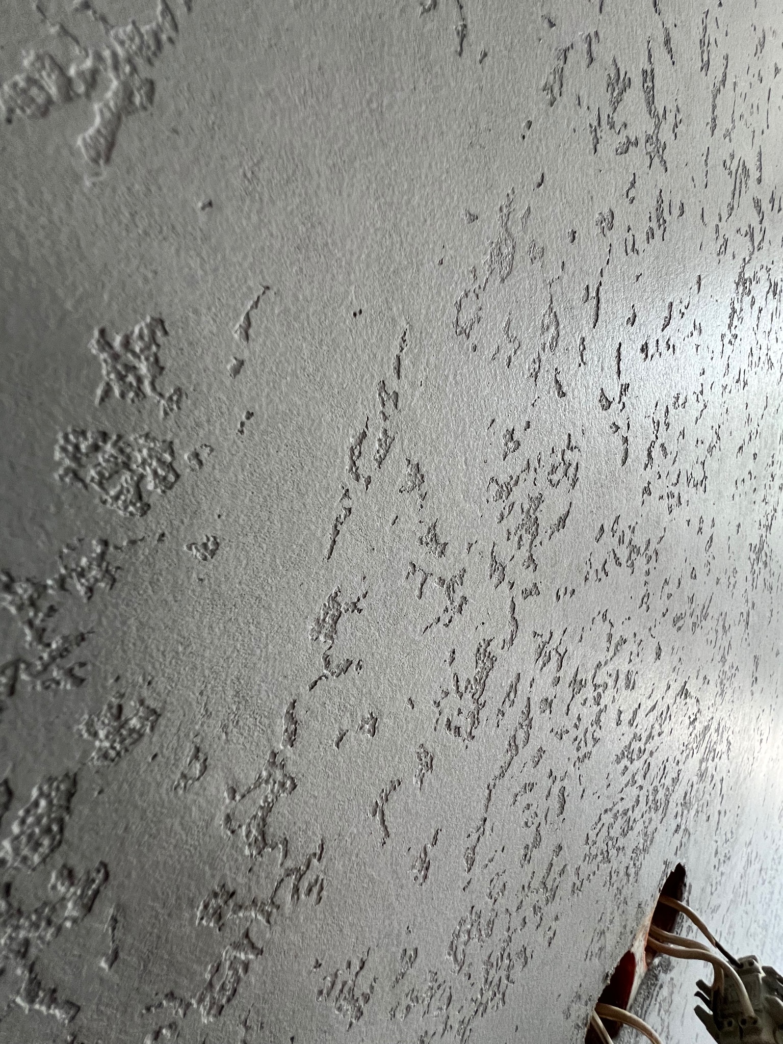 Szara ściana z dekoracyjną fakturą, widoczny otwór z przewodami elektrycznymi w dolnym rogu.