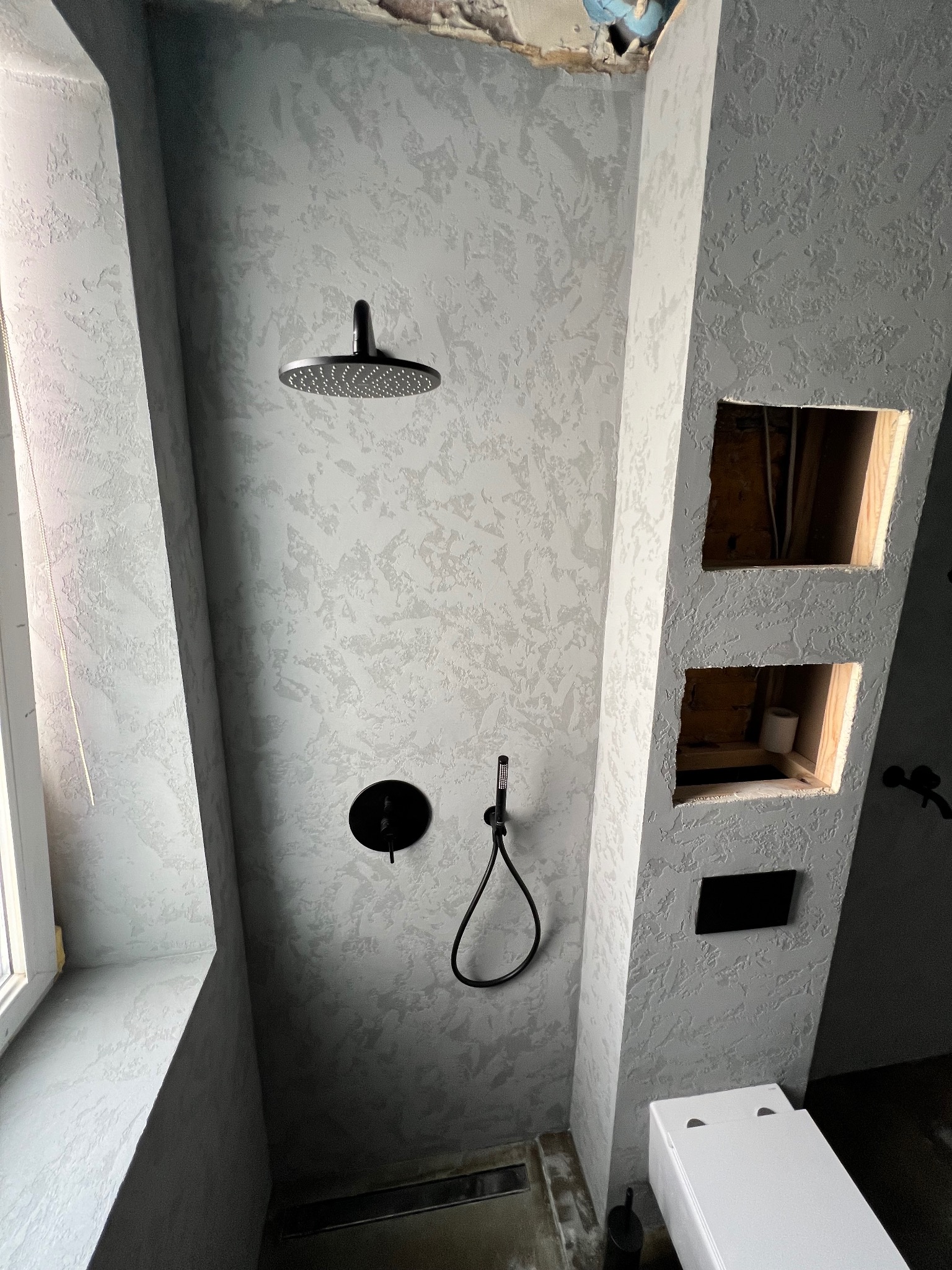 Nowoczesna łazienka w trakcie remontu: ściany pokryte szarą, dekoracyjną fakturą, czarna deszczownica i bateria, wnęki w ścianie w trakcie wykańczania, widoczne instalacje.