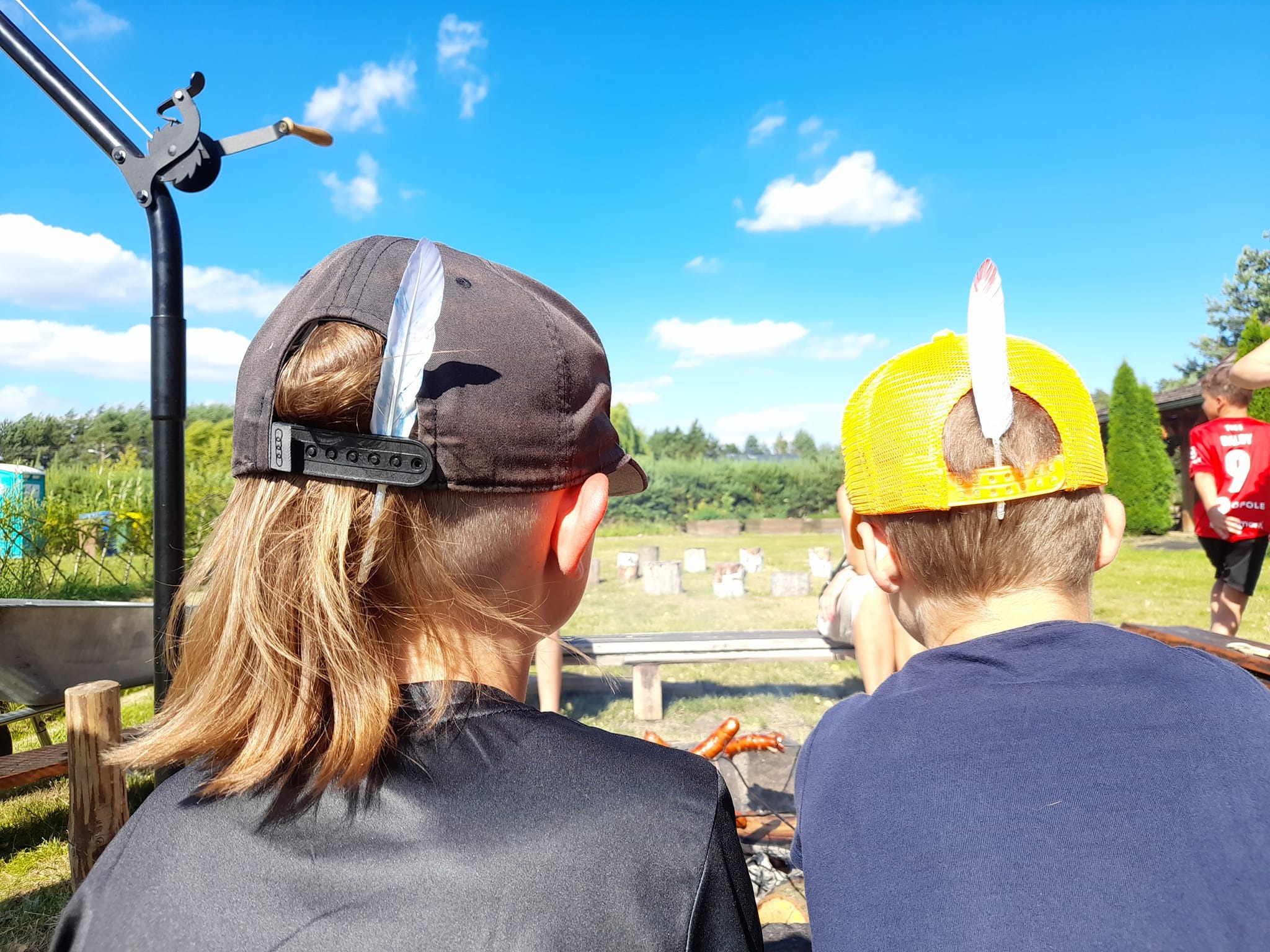 Chłopcy z piórami w czapkach siedzą przy ognisku, piekąc kiełbaski. W tle błękitne niebo i zieleń. Ujęcie z perspektywy osoby dorosłej, plenerowa impreza.