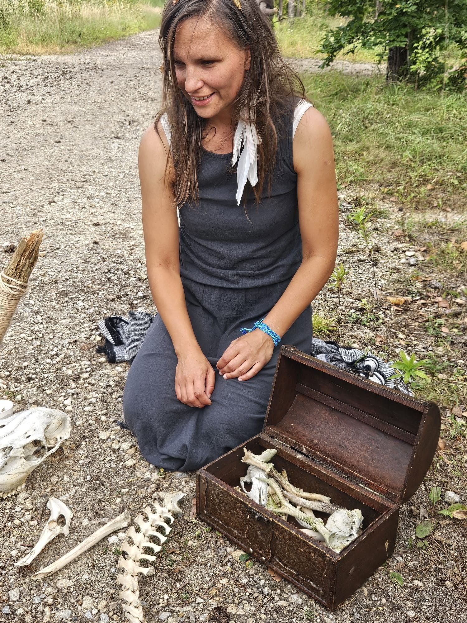 Kobieta w szarym kombinezonie klęczy obok drewnianej skrzyni z kośćmi, prawdopodobnie podczas gry terenowej z elementami edukacyjnymi na łonie natury.