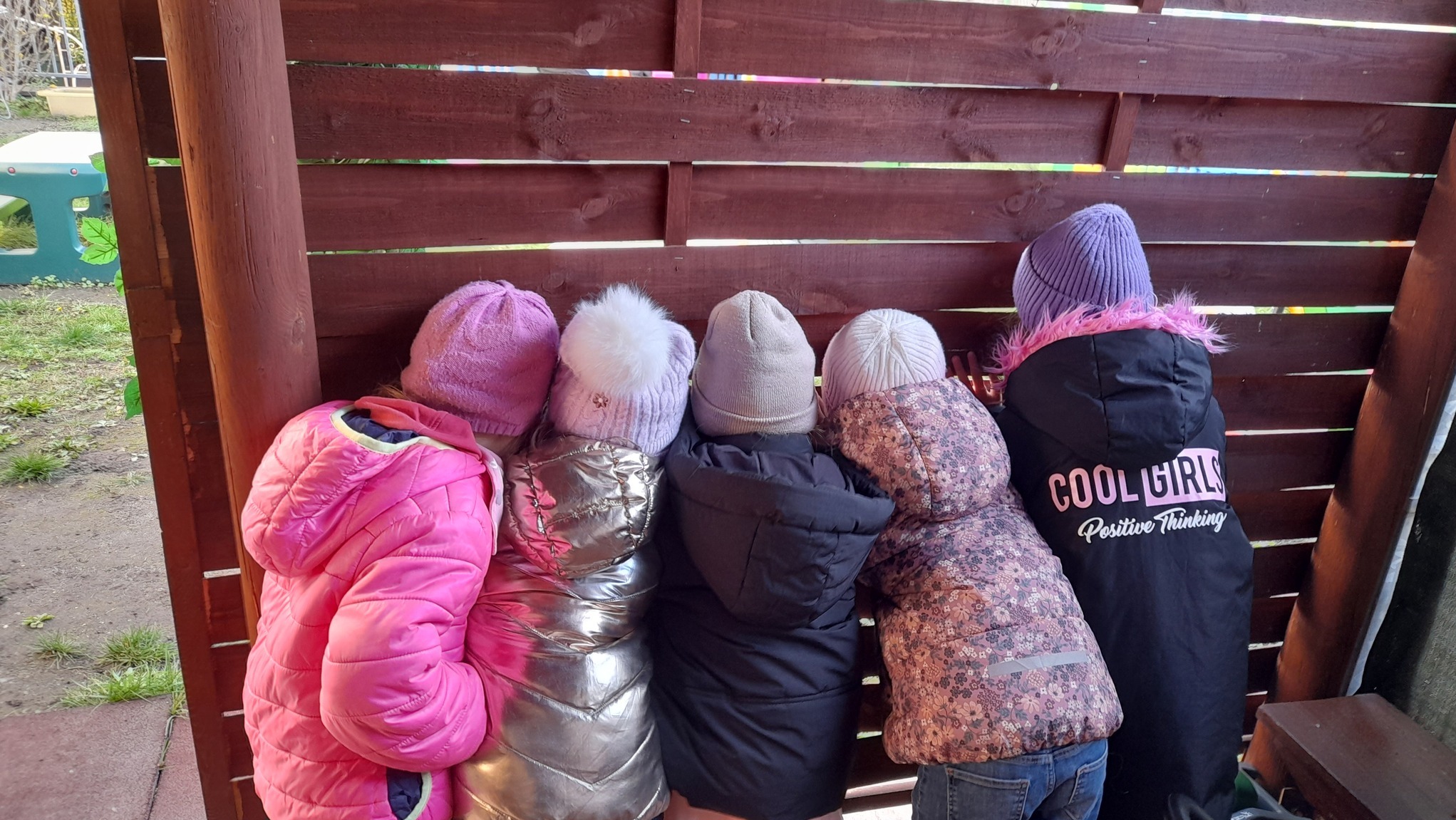 Pięcioro dzieci w kolorowych kurtkach i czapkach stoi obok drewnianej ściany, tyłem do widza. Na kurtce widoczny napis 'COOL GIRLS Positive Thinking'.