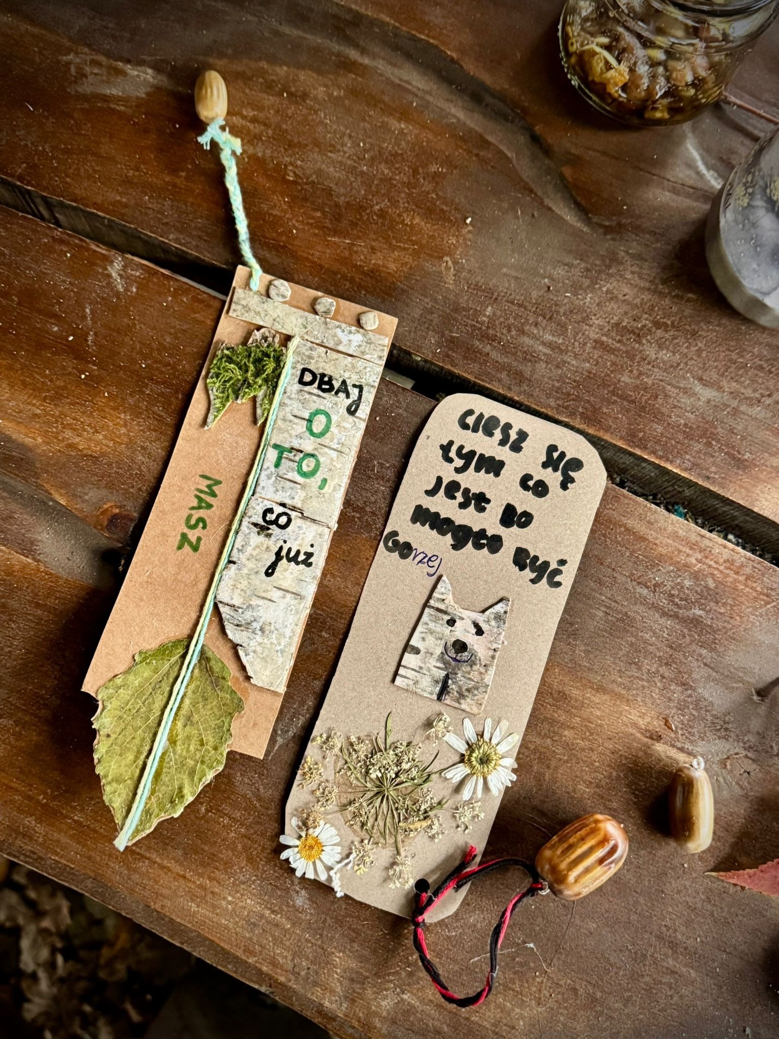 Ręcznie robione zakładki do książek z naturalnych materiałów: kory brzozy, suszonych kwiatów i liści, z napisami w języku polskim, leżące na drewnianym stole.