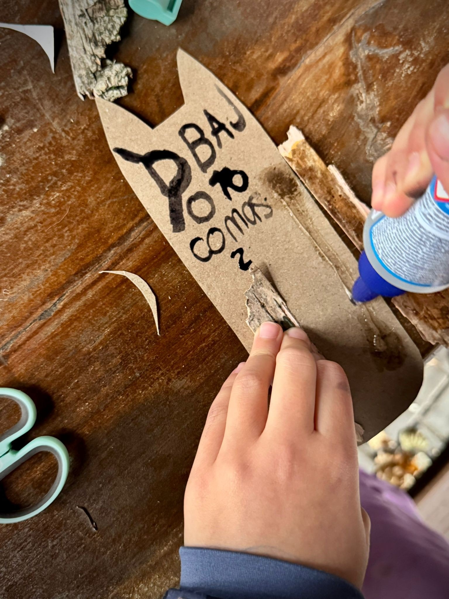 Dziecko tworzy ozdobę w kształcie zwierzęcia z kartonu i kawałków drewna, używając kleju. Widoczne nożyczki i inne elementy dekoracyjne na drewnianym stole.