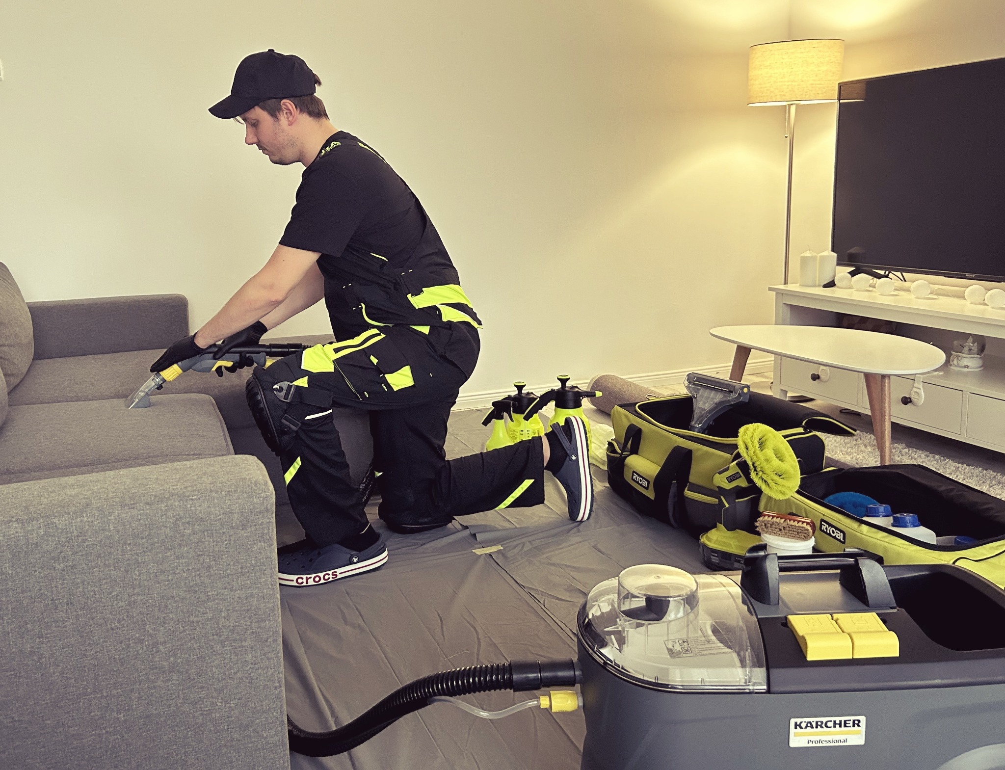 Mężczyzna w czarnym uniformie z żółtymi akcentami, klęczy na podłodze zabezpieczonej folią i czyści szarą kanapę za pomocą profesjonalnego odkurzacza Kärcher. W tle widoczny stolik kawowy...