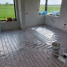 Instal-Tech - Instalacja ogrzewania podłogowego w surowym wnętrzu, widoczne rury grzewcze ułożone na folii z czerwoną siatką, wiadro z elementami instalacji obok, okno z widokiem na zielone pole w tle.