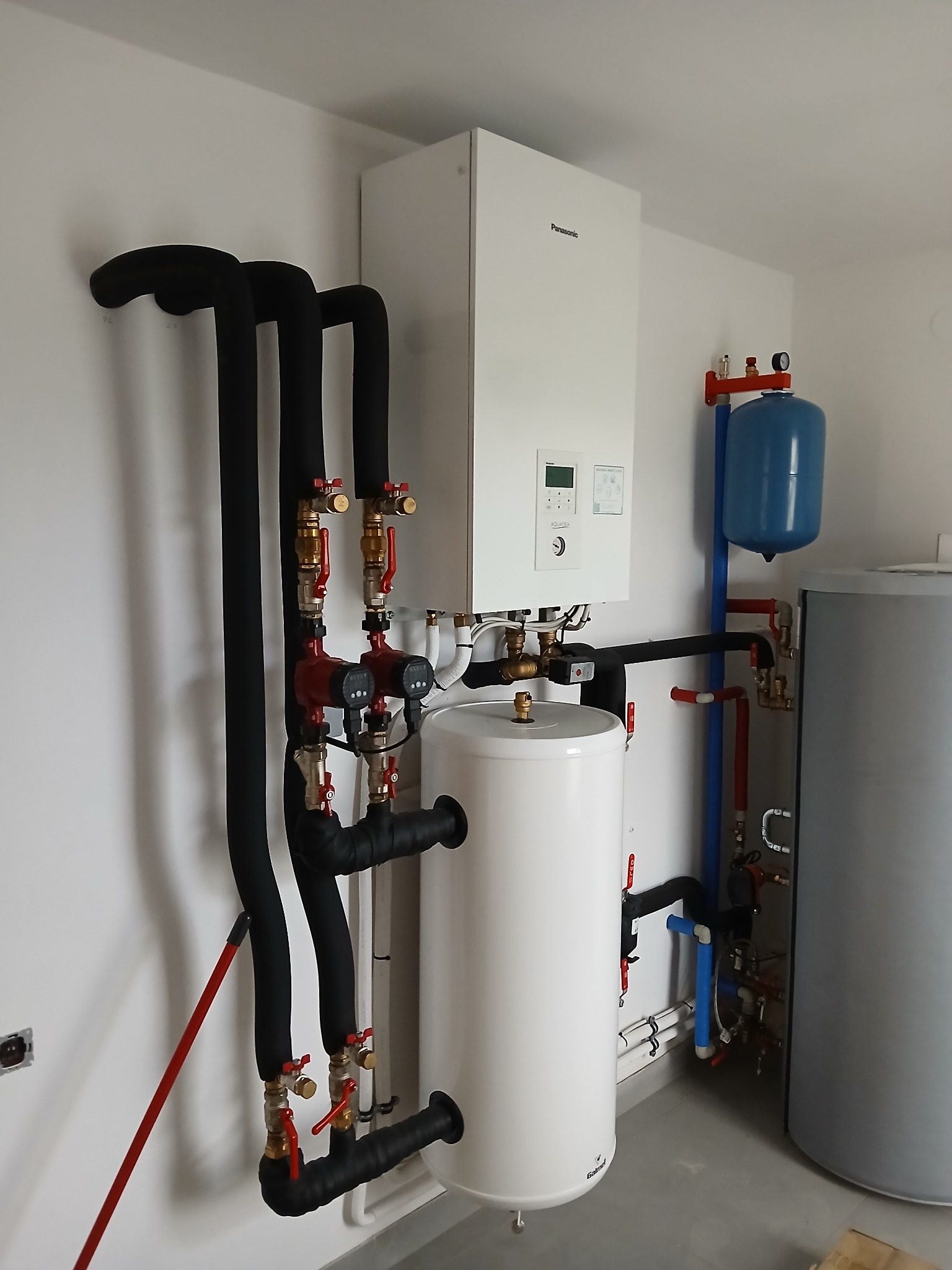 Nowoczesna kotłownia z piecem gazowym Panasonic, białym zasobnikiem ciepłej wody użytkowej, naczyniem przeponowym w kolorze niebieskim oraz rozbudowaną instalacją z czarnych rur izolowanych...