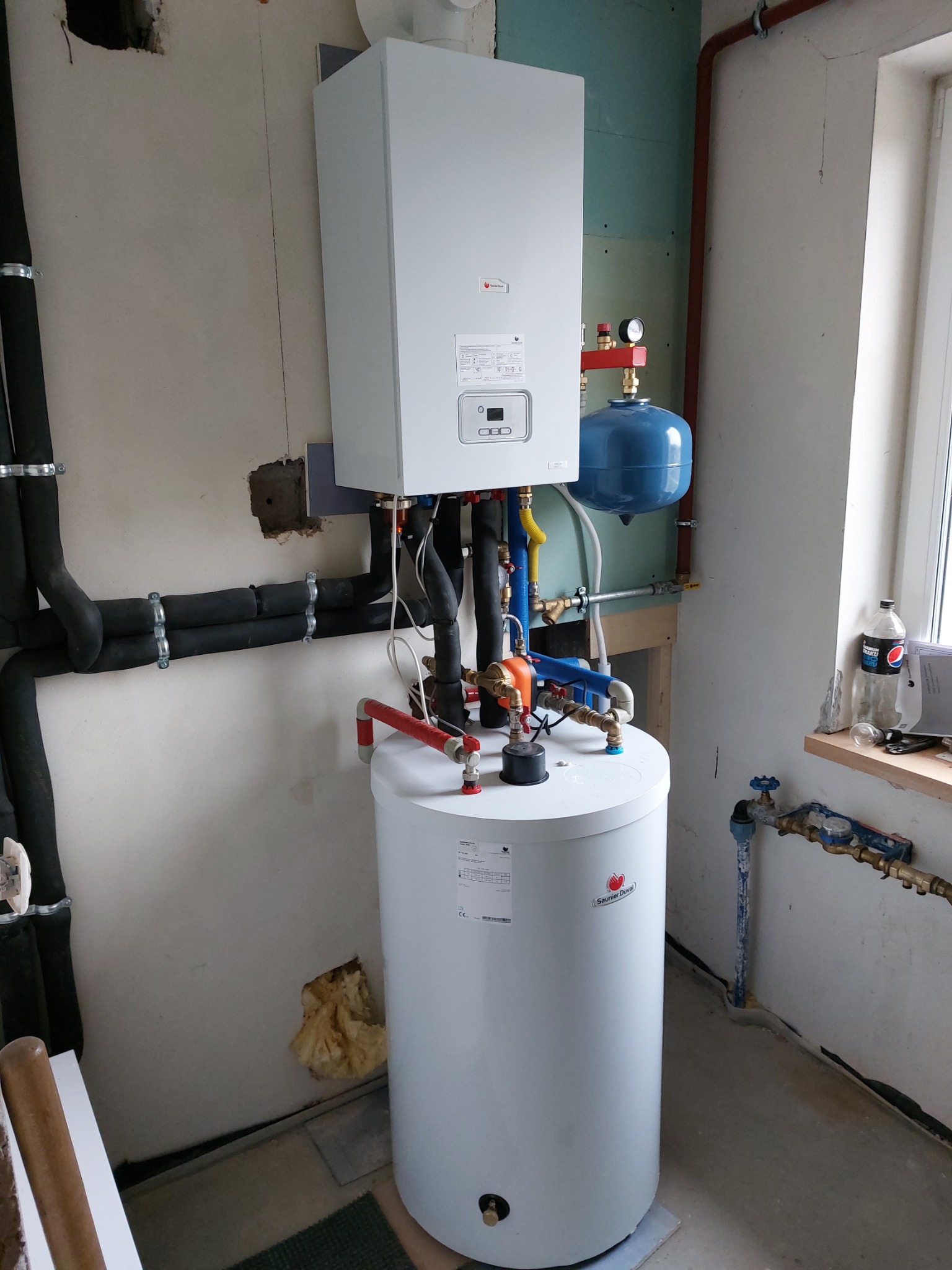 Biały piec gazowy kondensacyjny z podłączonym zasobnikiem ciepłej wody, zamontowany na ścianie w surowym pomieszczeniu z widocznymi rurami i elementami instalacji. Widoczne również czarne rury...