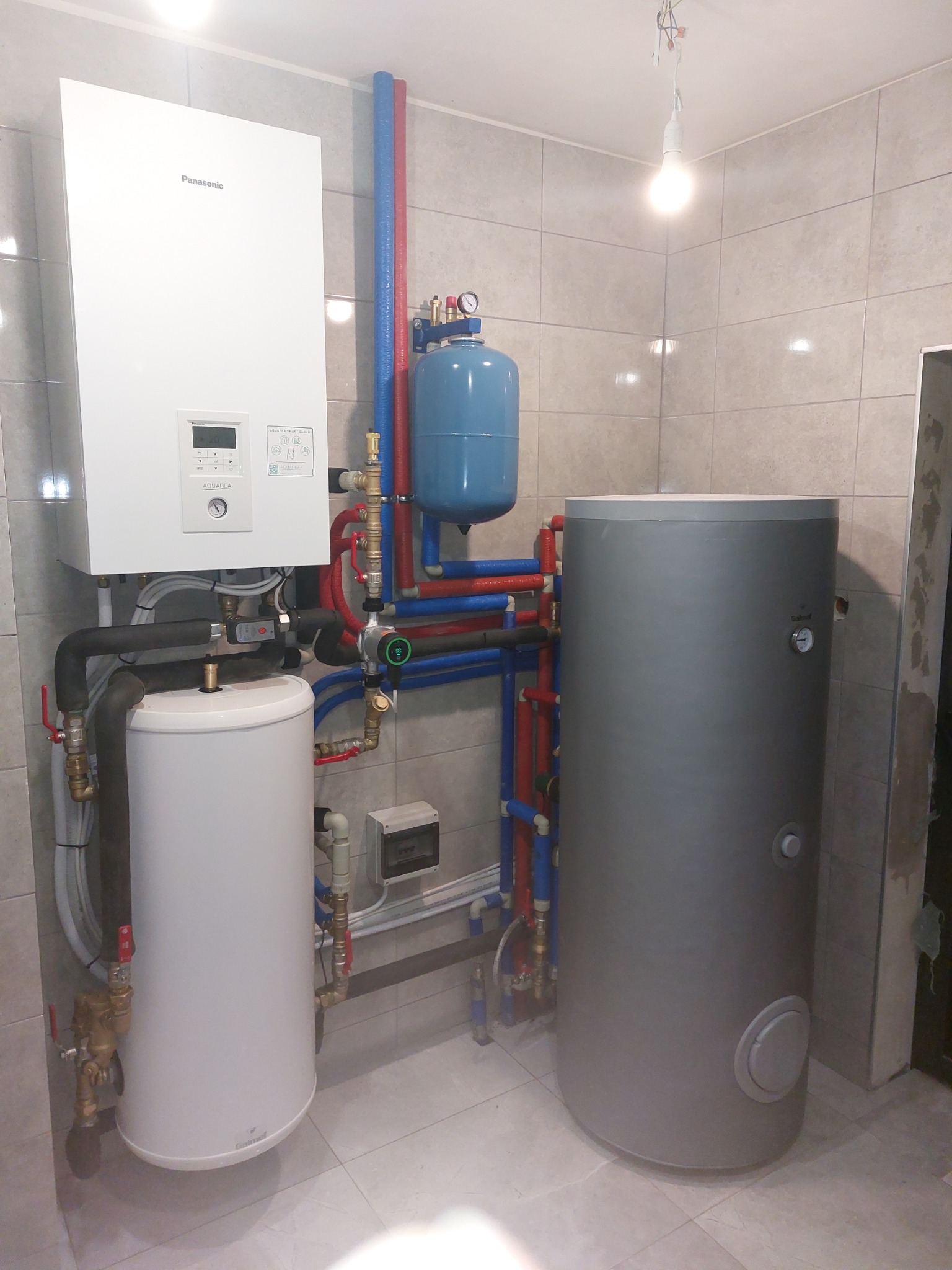 Biały piec gazowy Panasonic wiszący na ścianie, połączony rurami z niebieskim zbiornikiem wyrównawczym i dwoma zbiornikami na wodę (białym i szarym), instalacja w pomieszczeniu z jasnymi...