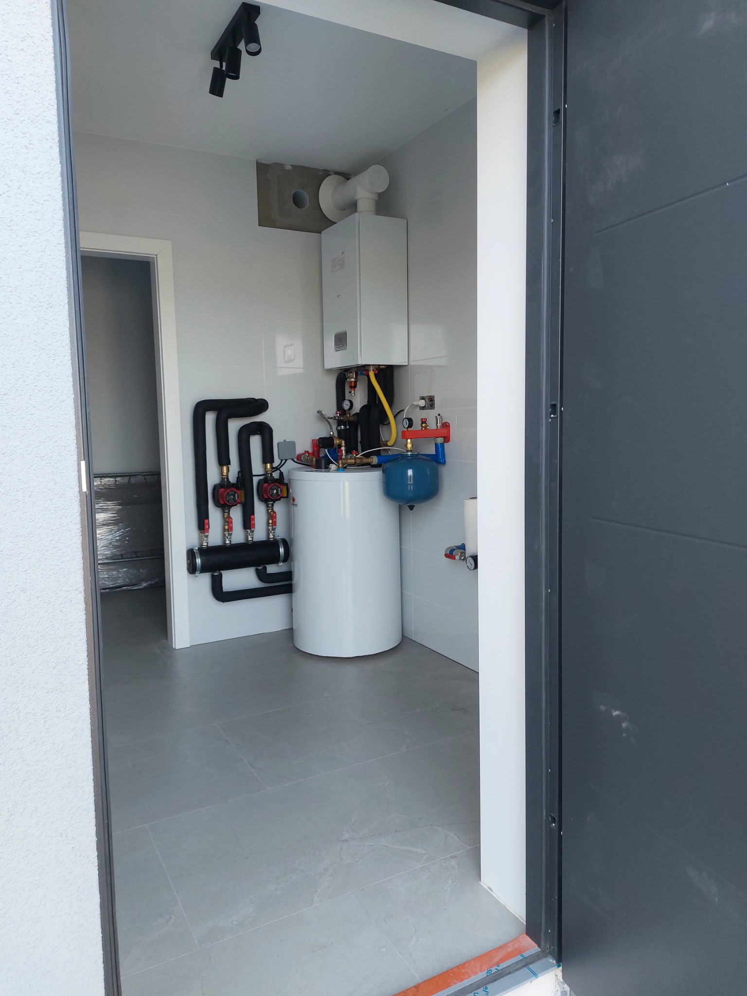 Wnętrze kotłowni z zamontowanym piecem gazowym, białym zasobnikiem ciepłej wody, grupą pompową i naczyniem przeponowym; widok zza otwartych drzwi, szare płytki podłogowe.
