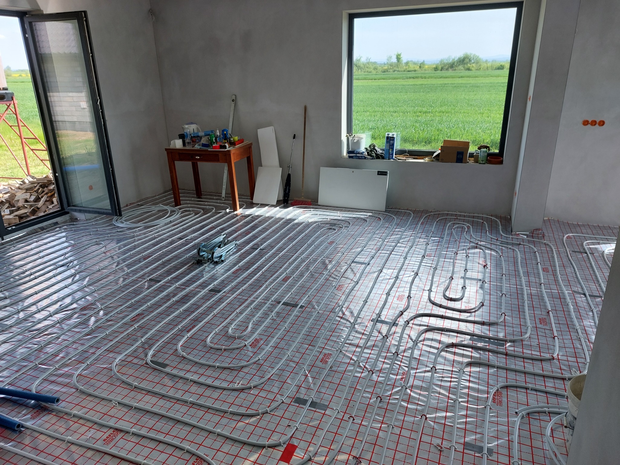 Rozłożone rury ogrzewania podłogowego na folii z czerwoną siatką w surowym wnętrzu, widok na zielone pole za oknem, narzędzia do montażu w tle.