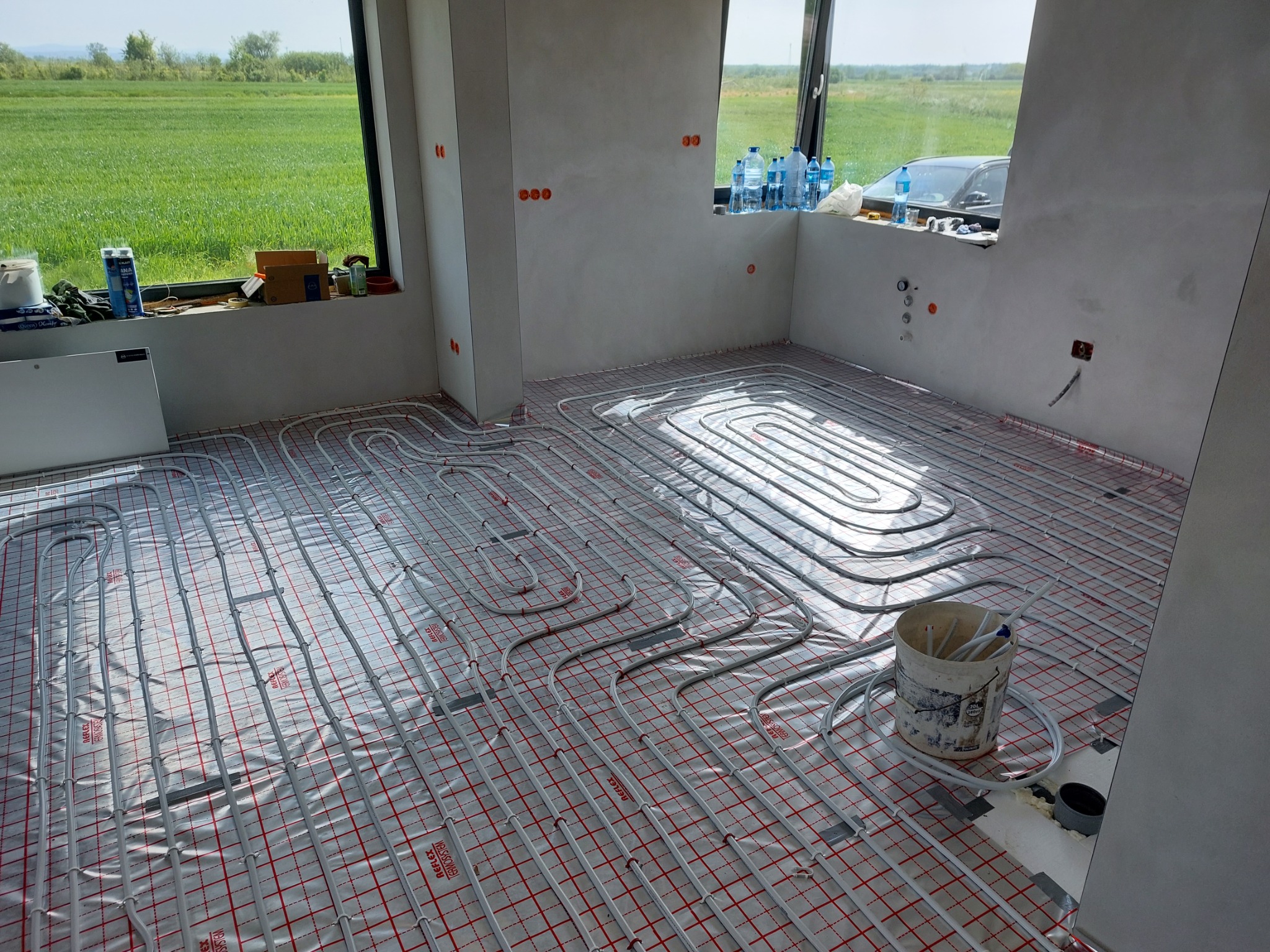 Instalacja ogrzewania podłogowego w surowym wnętrzu, widoczne rury grzewcze ułożone na folii z czerwoną siatką, wiadro z elementami instalacji obok, okno z widokiem na zielone pole w tle.