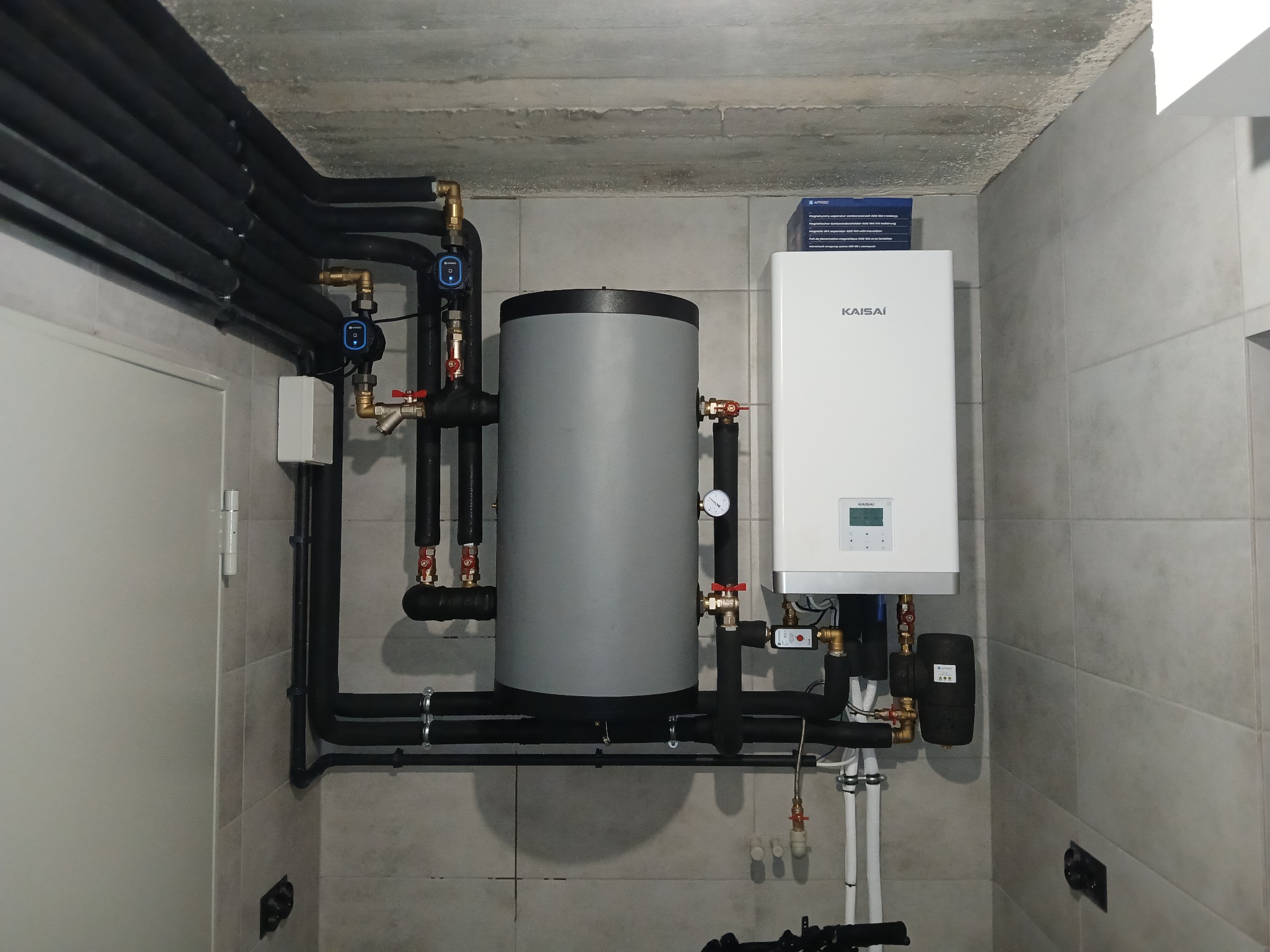 Kotłownia z szarym zasobnikiem ciepłej wody i białym piecem gazowym marki Kaisai, widoczne czarne rury instalacyjne z zaworami i manometrem, na ścianie płytki ceramiczne w odcieniach szarości.