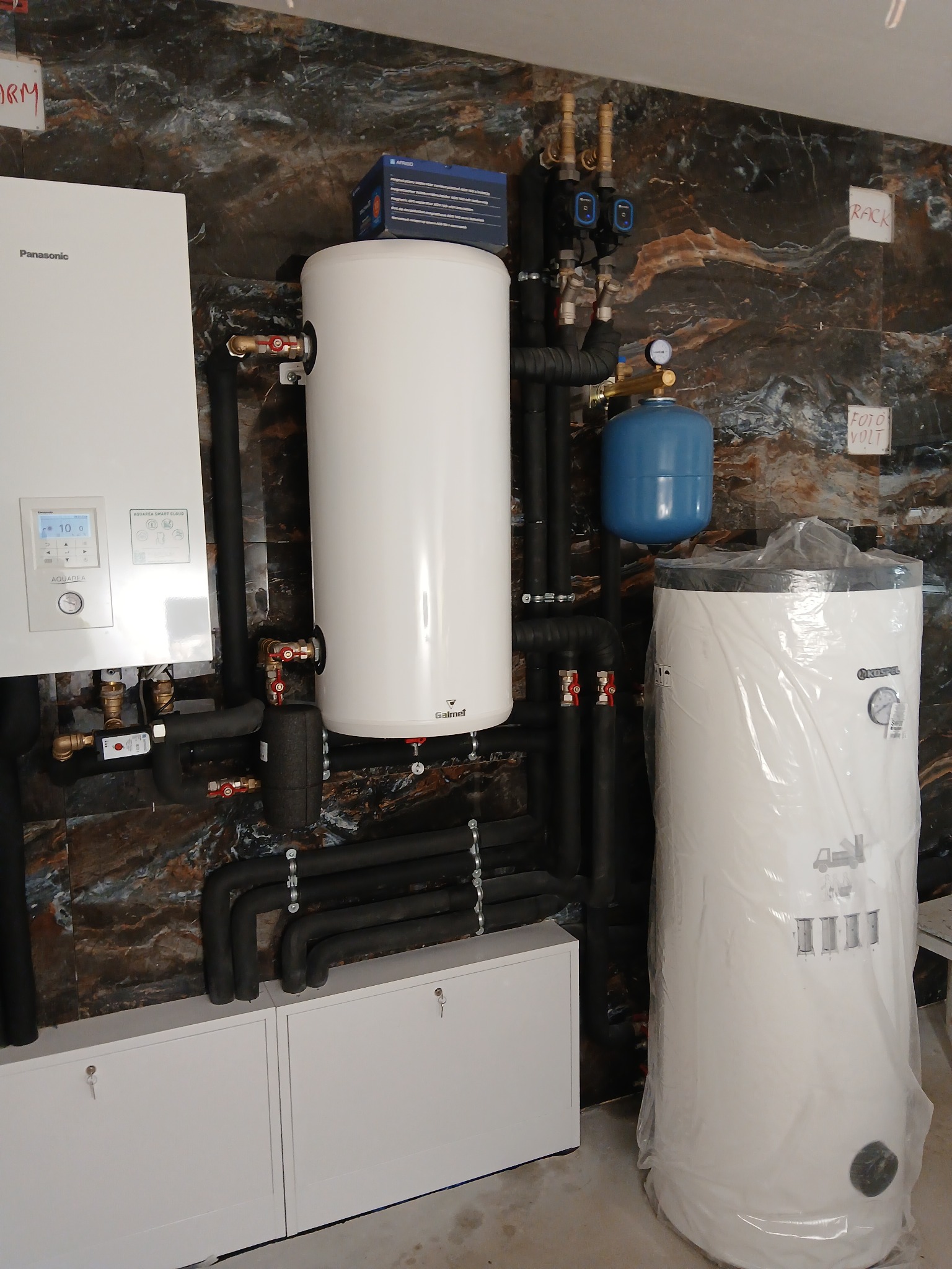 Zestaw instalacji grzewczej z piecem Panasonic Aquarea Smart Cloud, dwoma zbiornikami (jeden w folii) oraz elementami rur i zaworów na tle ściany z ciemnego marmuru.