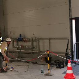 MARLIN DEEP SOLUTIONS Volodymyr Balandin - Hydraulicy w trakcie pracy przy instalacji wodnej w hali przemysłowej, widoczne rury, zawory, manometry, przewody, zabezpieczenia z łańcucha, w tle otwarte drzwi i zaparkowany bus