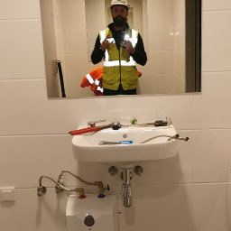 MARLIN DEEP SOLUTIONS Volodymyr Balandin - Instalacja umywalki z podgrzewaczem wody Blawar w łazience z białymi płytkami, widoczne narzędzia hydrauliczne i odbicie hydraulika w kamizelce w lustrze.