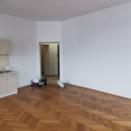 MARLIN DEEP SOLUTIONS Volodymyr Balandin - Wnętrze pokoju z parkietem ułożonym w jodełkę, białymi ścianami i sufitem, oraz wiszącą, okrągłą lampą z niebieskimi akcentami. Widoczne ślady po pracach remontowych: wiaderka, narzędzia i worki...