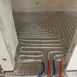 MARLIN DEEP SOLUTIONS Volodymyr Balandin - Rozłożona instalacja ogrzewania podłogowego w kształcie spirali, z widocznymi rurami w kolorze białym, czerwonym i niebieskim, przymocowanymi do folii z siatką.