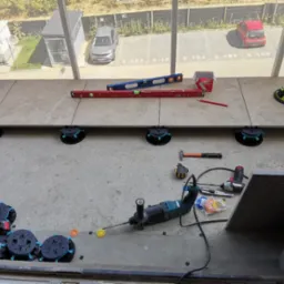 Montaż płytek na balkonie z użyciem poziomicy i podstawek regulacyjnych. Widoczne narzędzia i częściowo ułożona podłoga na tle parkingu.