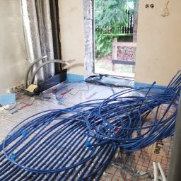MARLIN DEEP SOLUTIONS Volodymyr Balandin - Instalacja ogrzewania podłogowego w trakcie montażu. Widoczne niebieskie rury ułożone na folii izolacyjnej. W tle ściana z otwartym oknem na ogród.