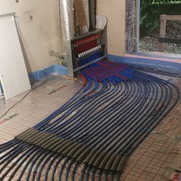 MARLIN DEEP SOLUTIONS Volodymyr Balandin - Rozłożona instalacja ogrzewania podłogowego z widocznym rozdzielaczem, rurami w kolorze niebieskim i czerwonym, oraz izolacją na części rur. Widok na okno w tle.