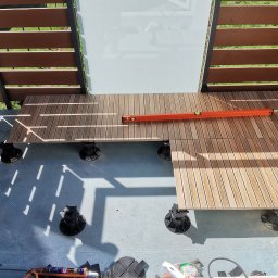 MARLIN DEEP SOLUTIONS Volodymyr Balandin - Montaż tarasu drewnianego na regulowanych wspornikach, poziomowany poziomicą, widok z góry na balkon z drewnianą zabudową i fragmentem ogrodzenia.