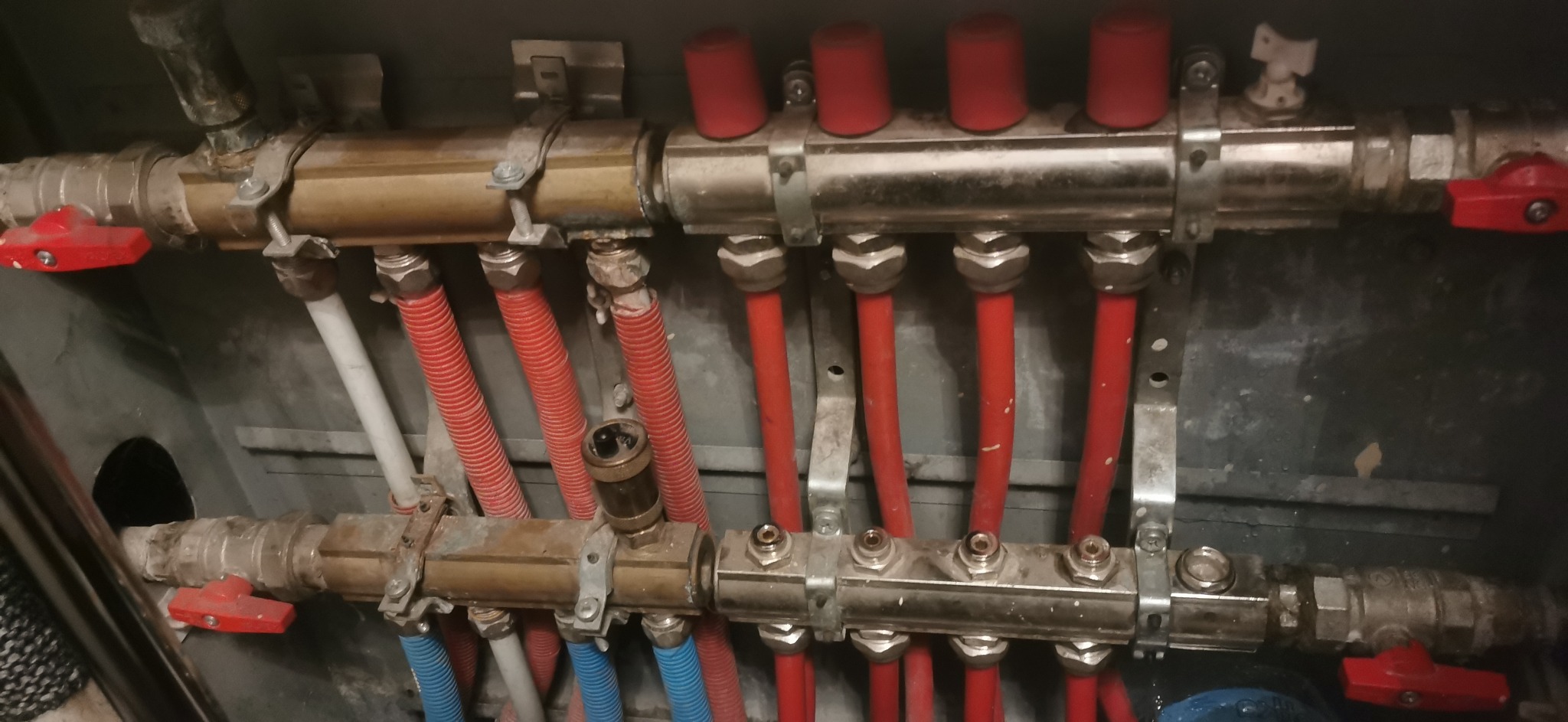 Rozdzielacz instalacji grzewczej z czerwonymi i niebieskimi rurami, widoczne zawory z czerwonymi uchwytami, mocowanie na metalowych wspornikach do ściany.