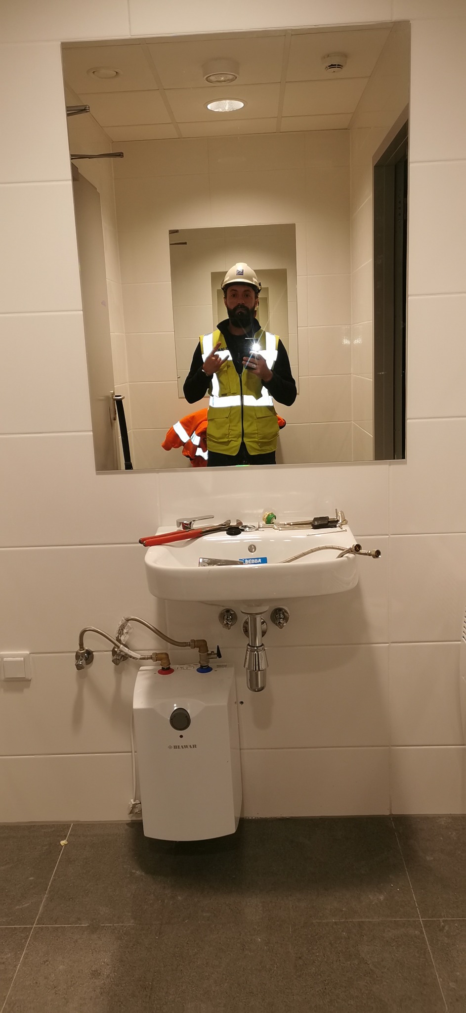 Instalacja umywalki z podgrzewaczem wody Blawar w łazience z białymi płytkami, widoczne narzędzia hydrauliczne i odbicie hydraulika w kamizelce w lustrze.