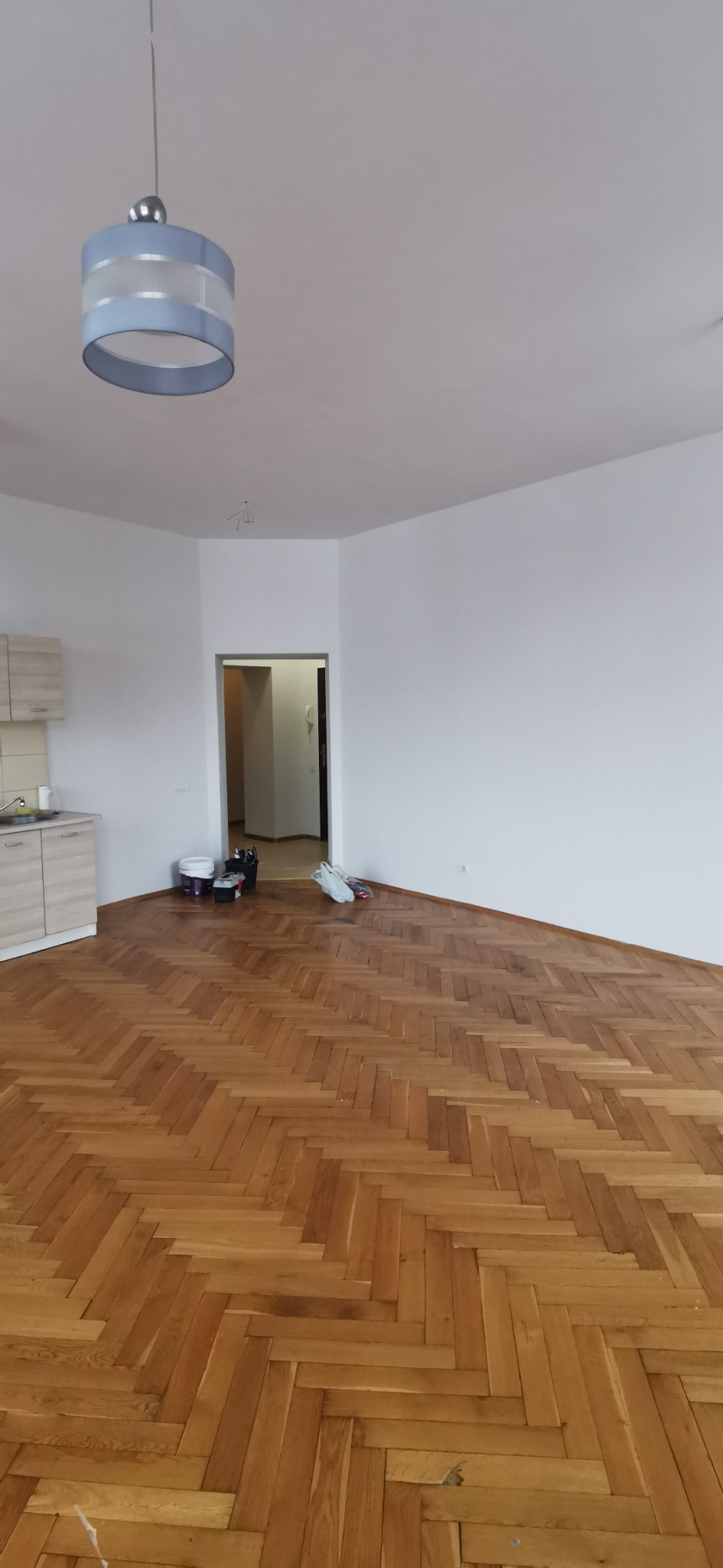 Wnętrze pokoju z parkietem ułożonym w jodełkę, białymi ścianami i sufitem, oraz wiszącą, okrągłą lampą z niebieskimi akcentami. Widoczne ślady po pracach remontowych: wiaderka, narzędzia i worki...