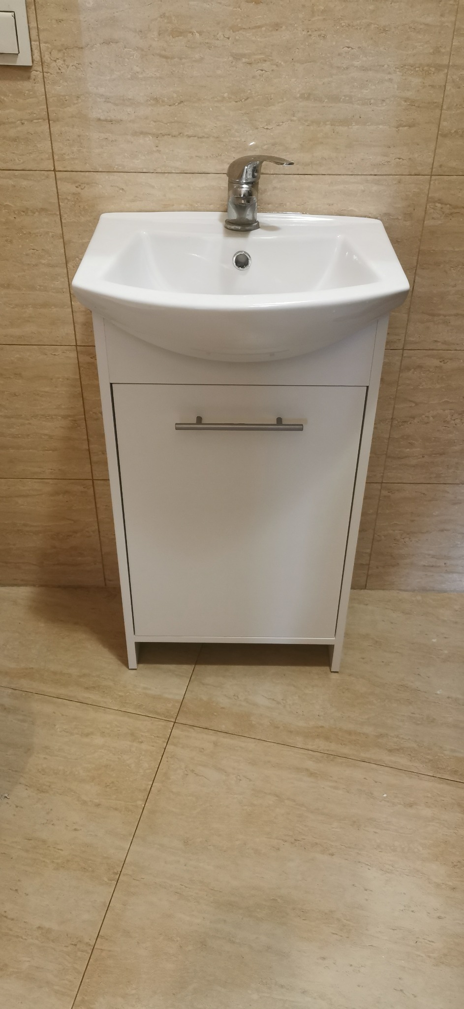 Biała umywalka z szafką i chromowaną baterią, zamontowana na tle beżowych płytek ściennych i podłogowych.