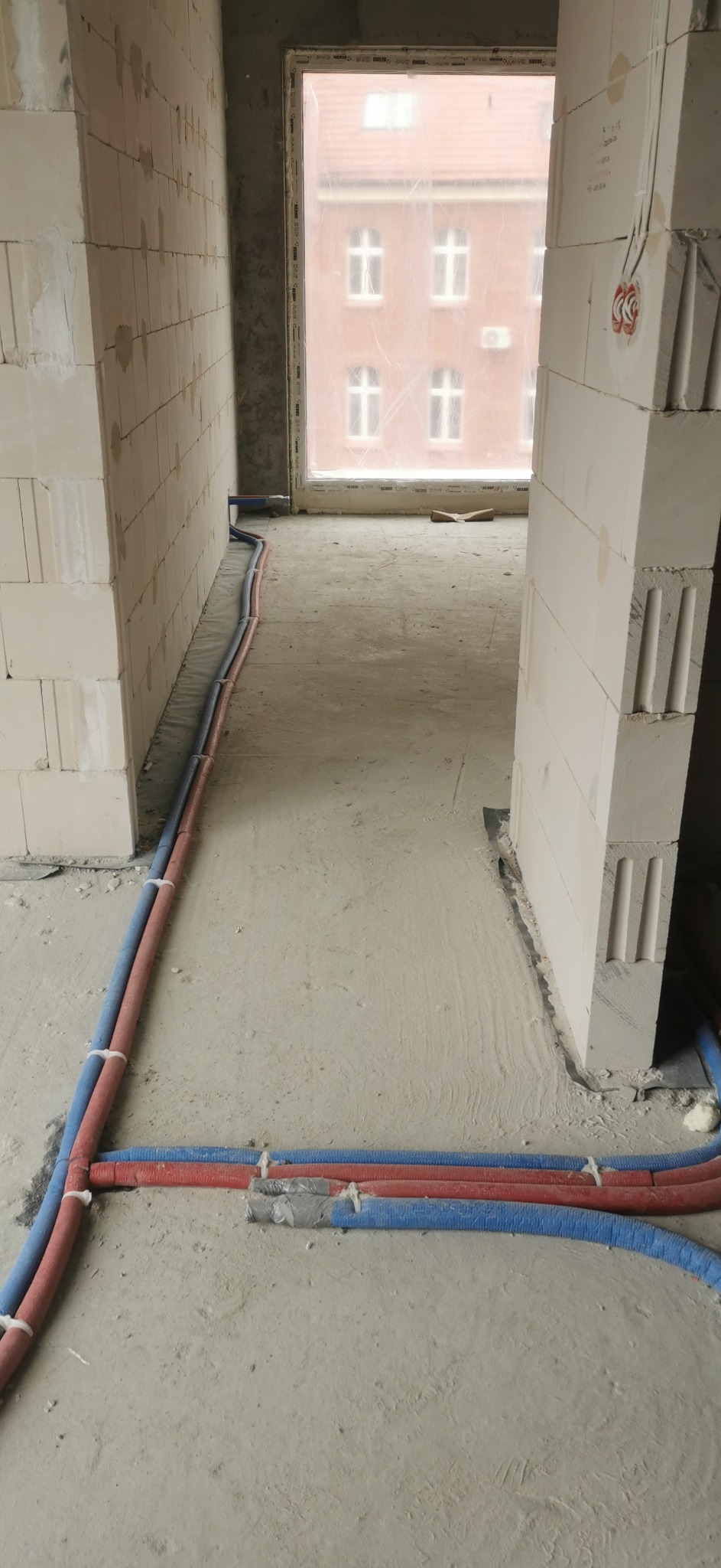 Wewnętrzne ułożenie rur grzewczych (czerwone i niebieskie) w surowym stanie, widoczne łączenia zabezpieczone taśmą, ściany z białych bloczków, w tle okno z widokiem na budynek.