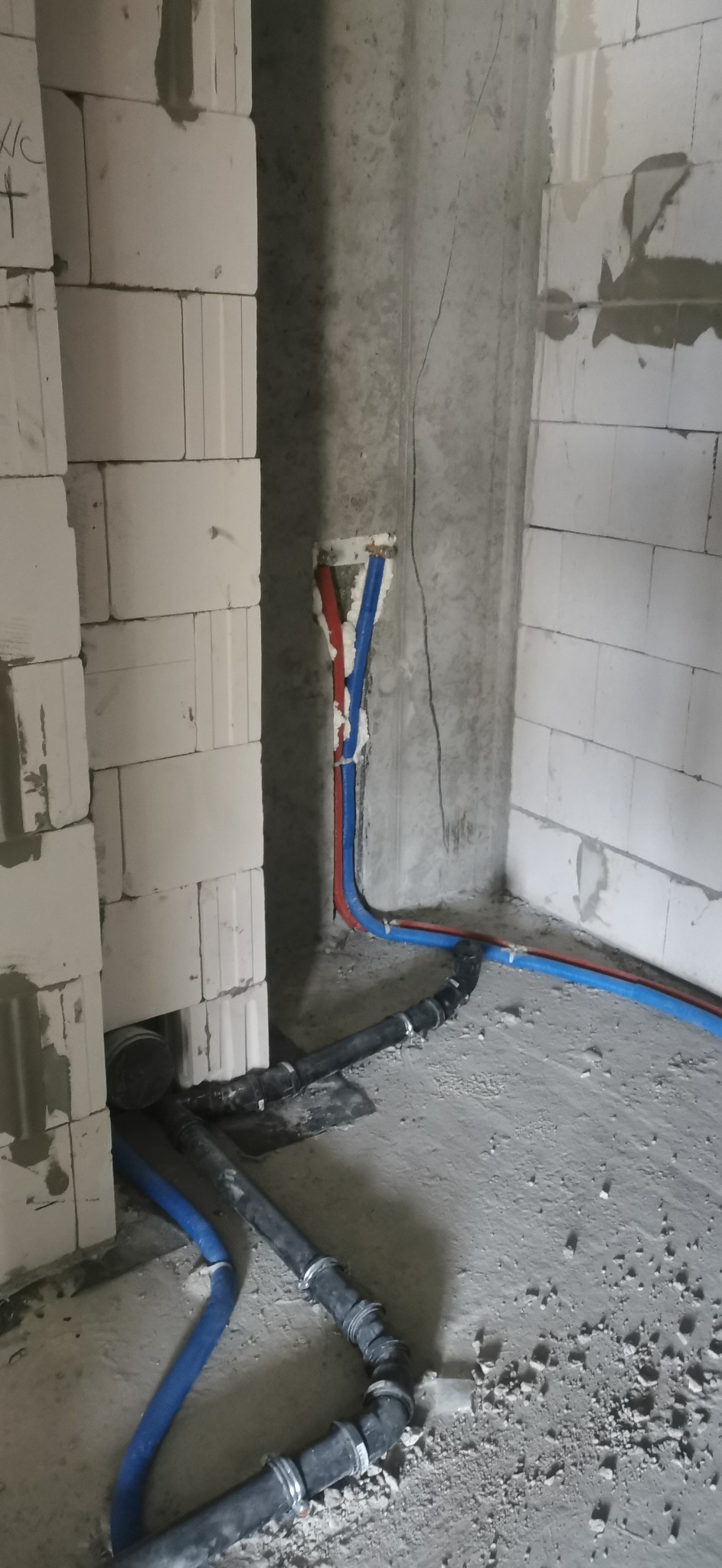 Wnętrze w trakcie prac instalacyjnych: widoczne rury doprowadzające wodę (czerwona i niebieska) oraz czarne rury kanalizacyjne z licznymi złączkami, prowadzone po surowym betonowym podłożu...