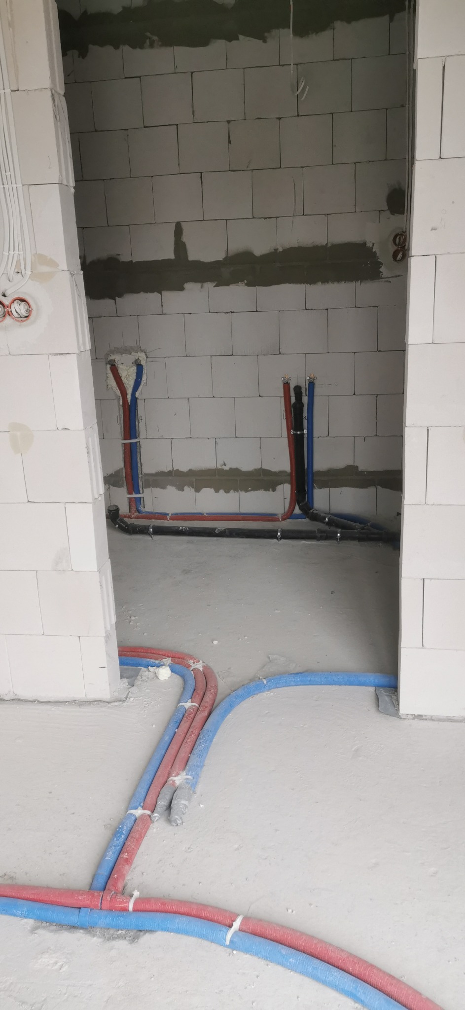 Surowe wnętrze z instalacją hydrauliczną i grzewczą w trakcie montażu, widoczne rury w kolorach niebieskim i czerwonym przymocowane do ścian z bloczków betonowych oraz prowadzone po podłodze...