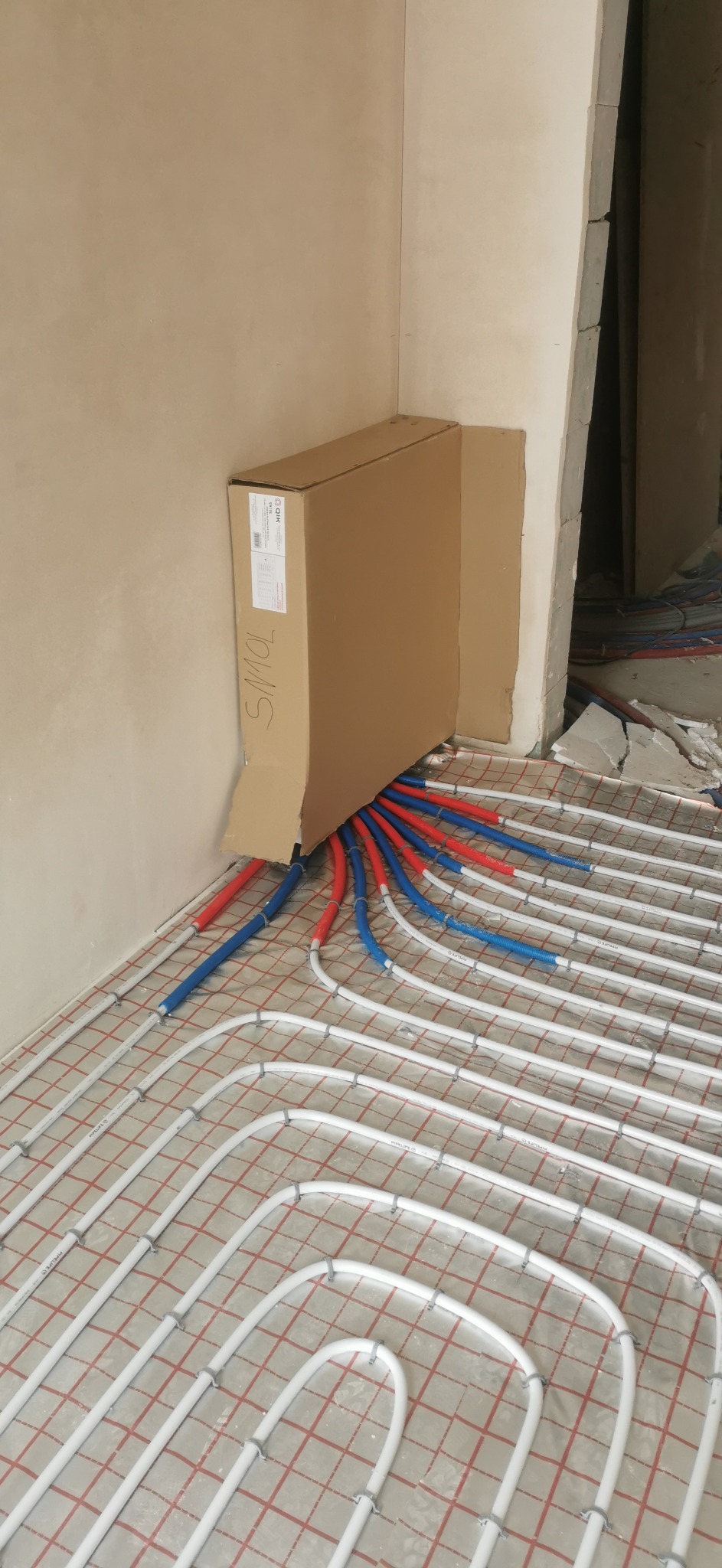 Białe rury ogrzewania podłogowego rozłożone na czerwonej macie, z wystającymi rurami doprowadzającymi w kolorach niebieskim i czerwonym, przy ścianie i kartonie.