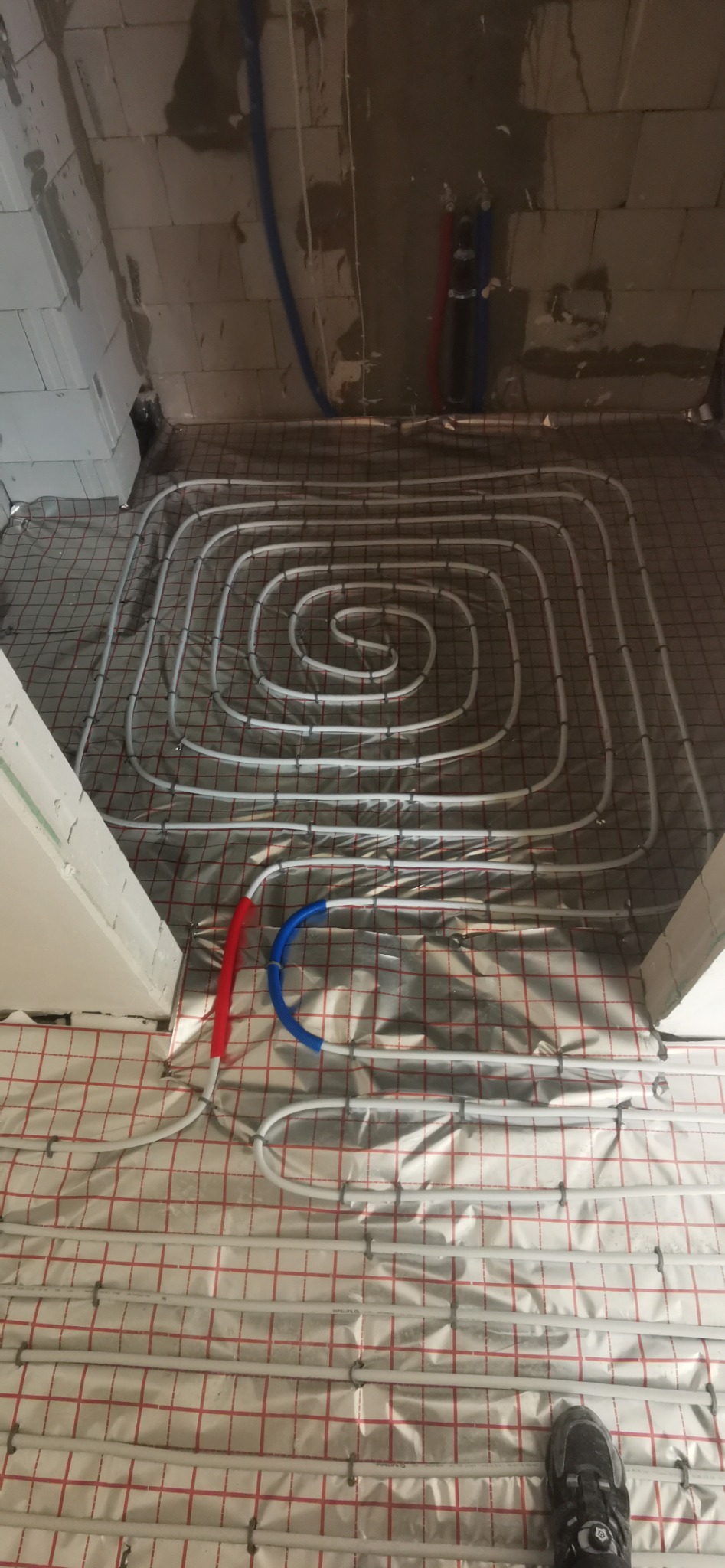 Spiralnie ułożone białe rury ogrzewania podłogowego na srebrnej folii z czerwoną siatką, widoczne elementy instalacji na ścianie i fragment buta.