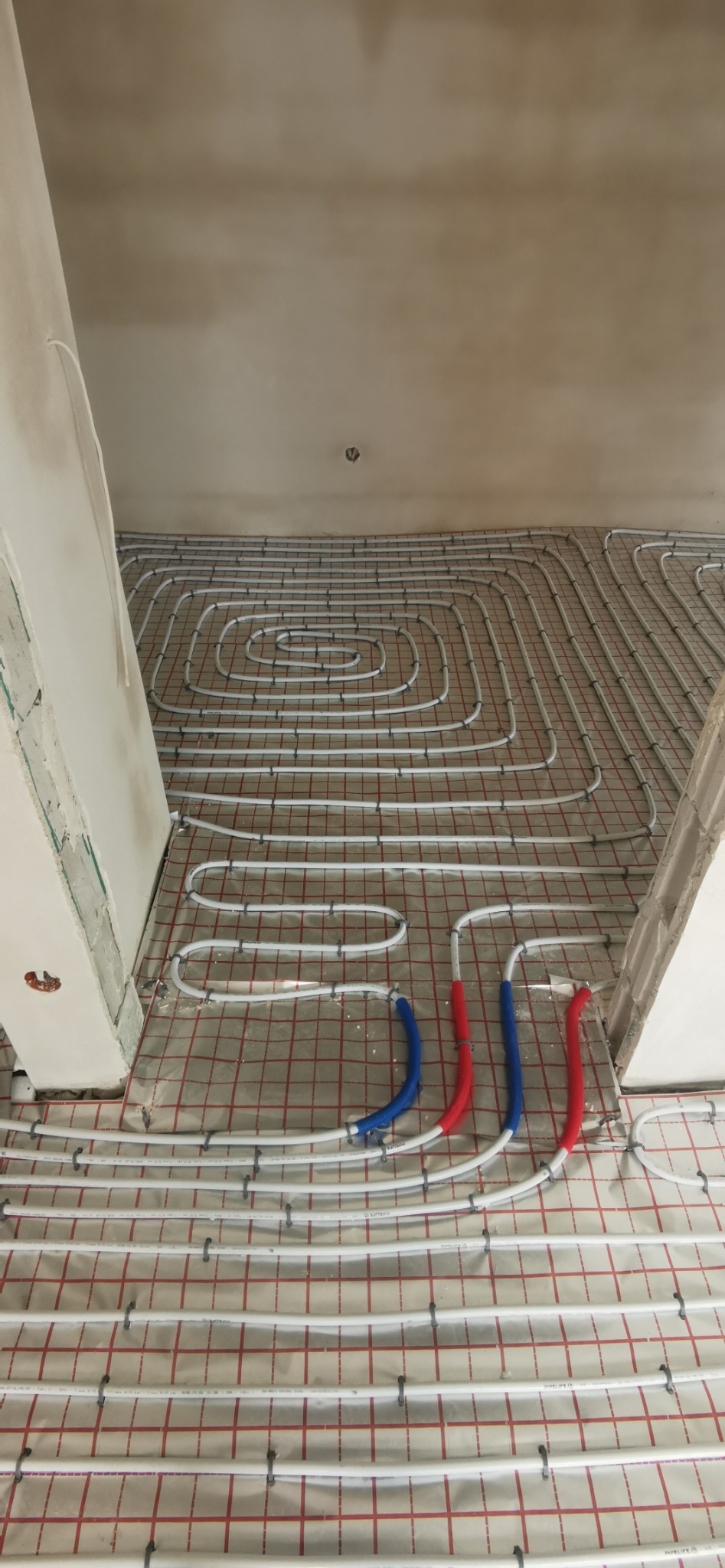 Rozłożona instalacja ogrzewania podłogowego w kształcie spirali, z widocznymi rurami w kolorze białym, czerwonym i niebieskim, przymocowanymi do folii z siatką.