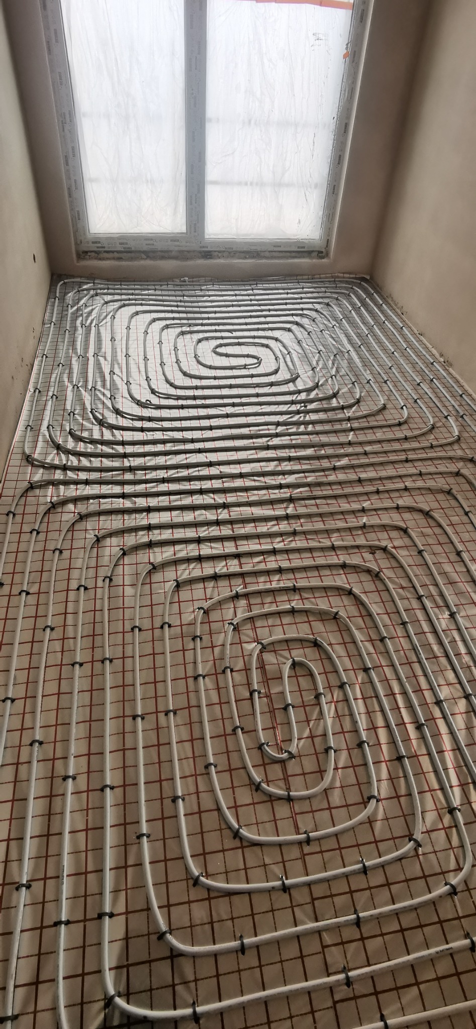 Białe rury ogrzewania podłogowego ułożone spiralnie na izolacji z czerwoną kratką, widok z góry, okno dachowe w tle.