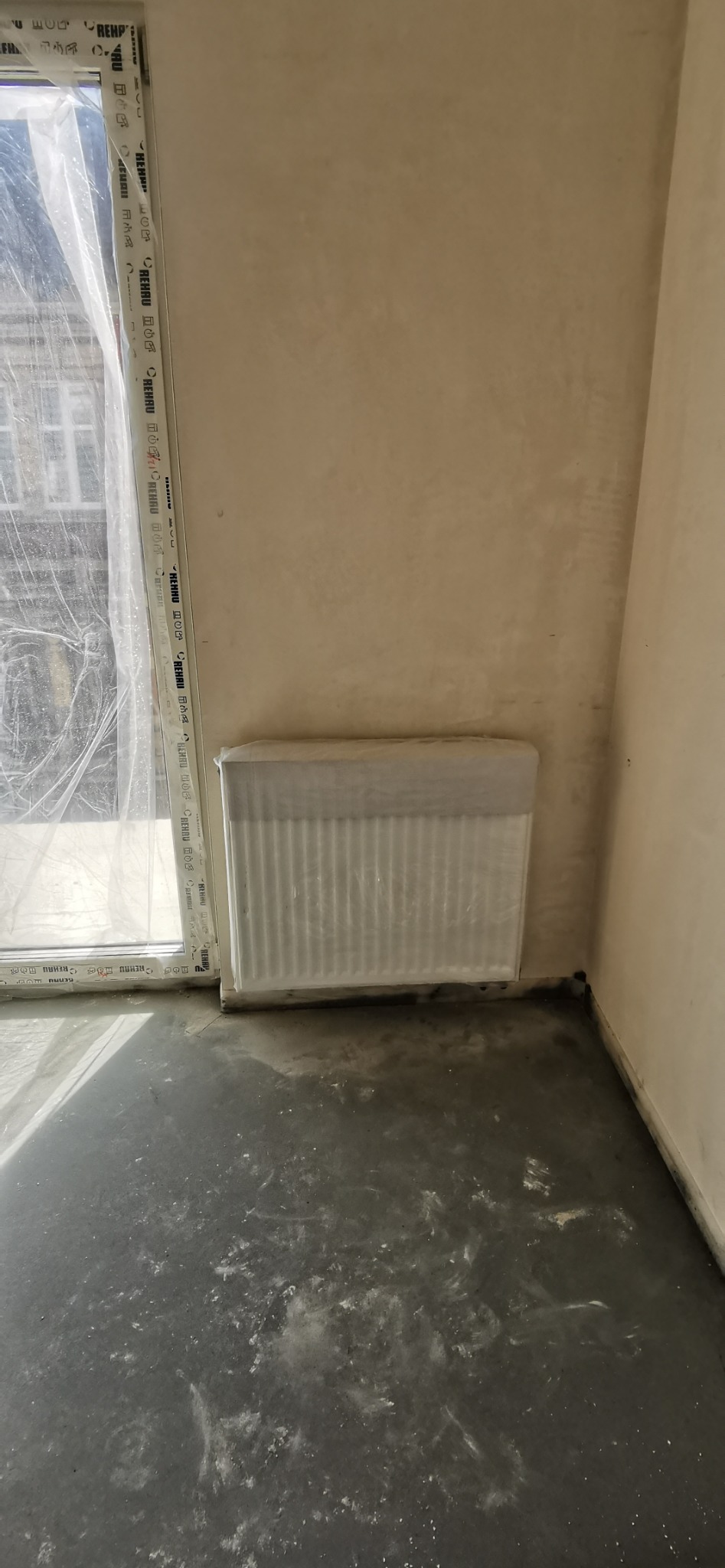 Ogrzejnik zakryty folią ochronną podczas prac wykończeniowych. Widoczna ściana po gruntowaniu i okno z folią. Podłoga częściowo pokryta pyłem.