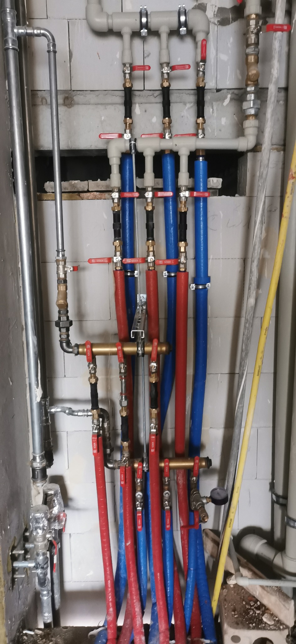 Złożona instalacja hydrauliczna z rurami w kolorach czerwonym i niebieskim, zaworami i elementami mocującymi, zamontowana na tle surowej ściany z pustaków.