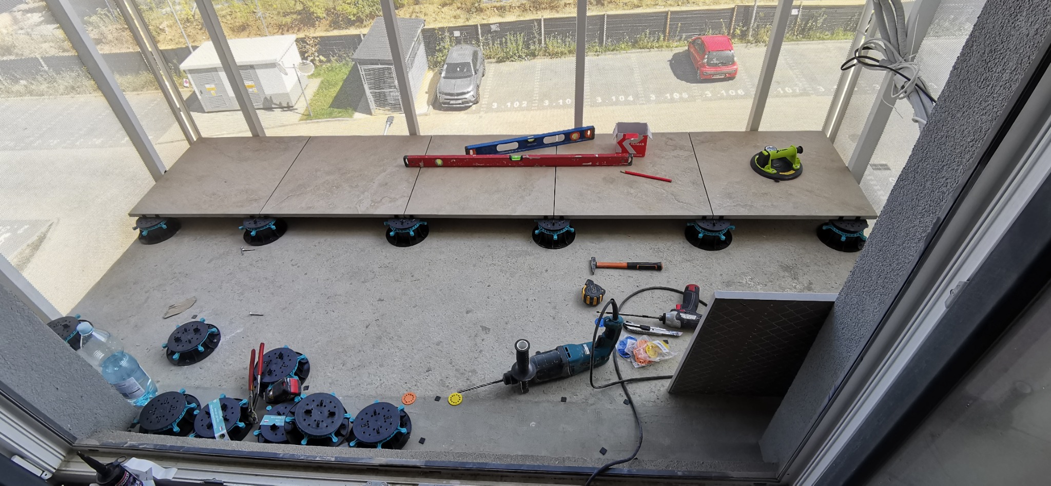 Montaż płytek na balkonie z użyciem poziomicy i podstawek regulacyjnych. Widoczne narzędzia i częściowo ułożona podłoga na tle parkingu.
