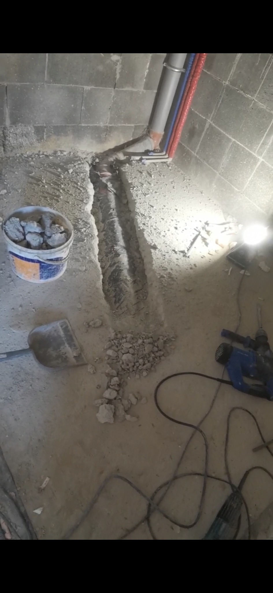 Wykopany rów w podłodze pod instalację hydrauliczną, widoczne rury, gruz, wiadro z resztkami betonu, szpadel i wiertarka udarowa. Prace w toku, surowe wykończenie.